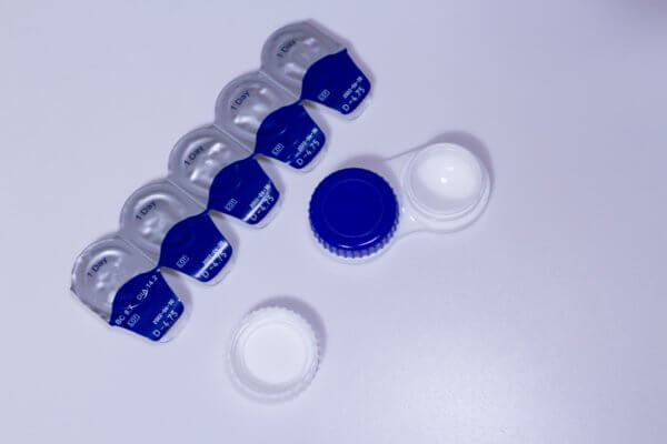 This image shows a contact lens case and several single-use contact lens solution packets arranged on a white surface. The contact lens case is open, with one lid removed, and the solution packets are lined up nearby.