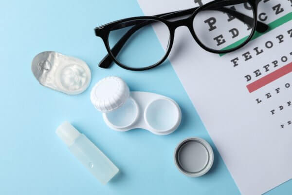 The image shows a pair of black-rimmed eyeglasses, a white contact lens case with its cap removed, an open blister pack with a contact lens, a small bottle of contact lens solution, and part of an eye chart with letters of varying sizes.