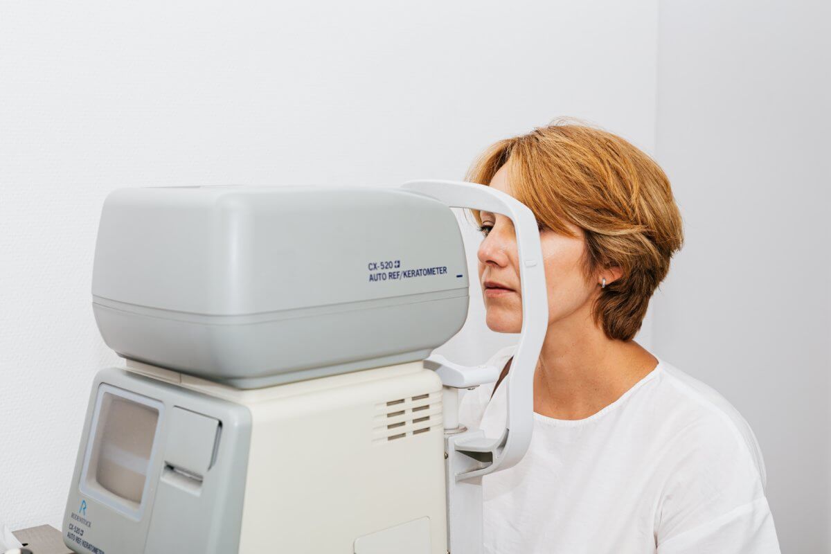 This image shows a woman undergoing an eye examination with an auto-refractor/keratometer machine, commonly used in optometry to measure the curvature of the eye's surface and assess vision needs.