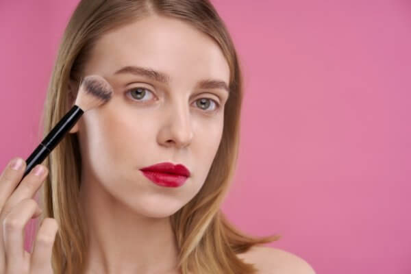 A young woman applies makeup with a brush to her cheek while looking at the camera. She has red lipstick and is set against a pink background.