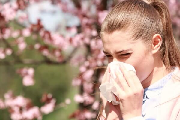  This image shows a young woman sneezing into a tissue, likely due to seasonal allergies, with a background of pink blossom trees, suggesting it's springtime.